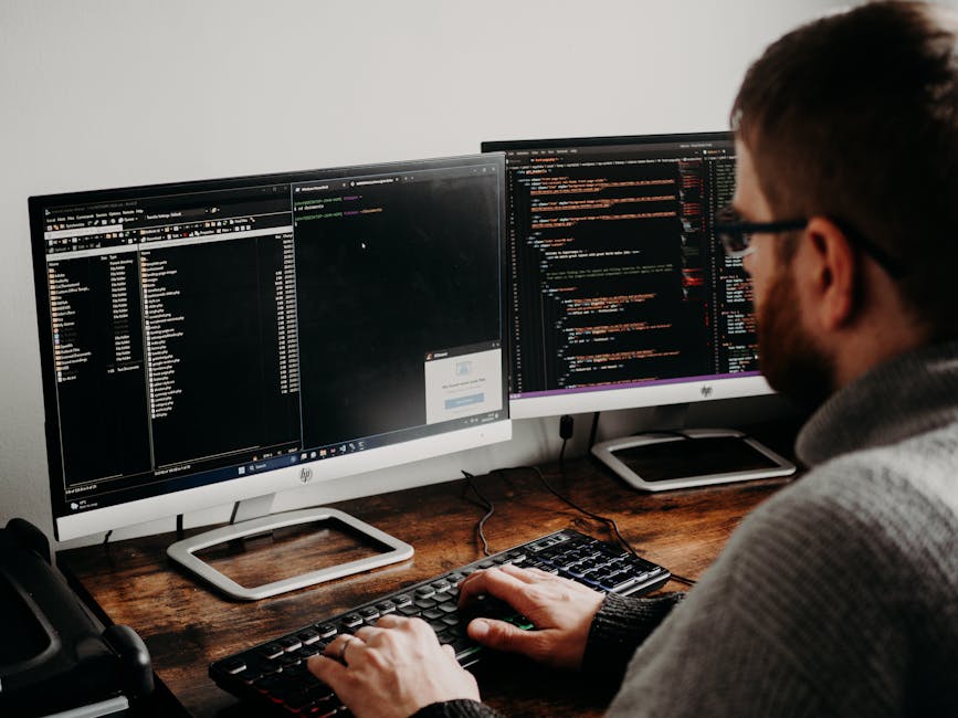 A person with short brown hair, wearing glasses and a grey sweater, working at a desk with two large computer monitors displaying lines of code with syntax highlighting in dark mode. The workspace fea